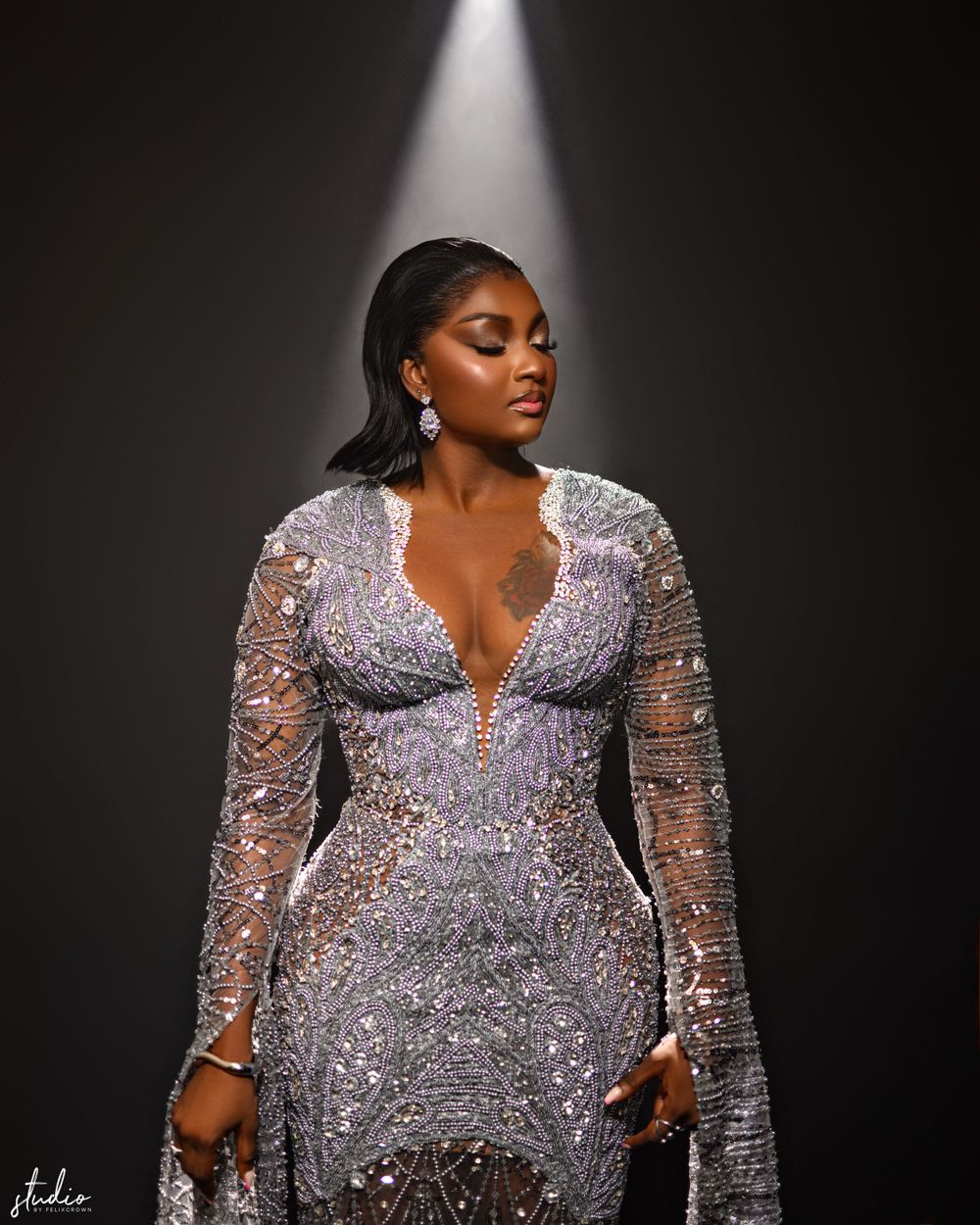 A figure in a stunning silver beaded gown with long sheer sleeves poses dramatically under a spotlight.