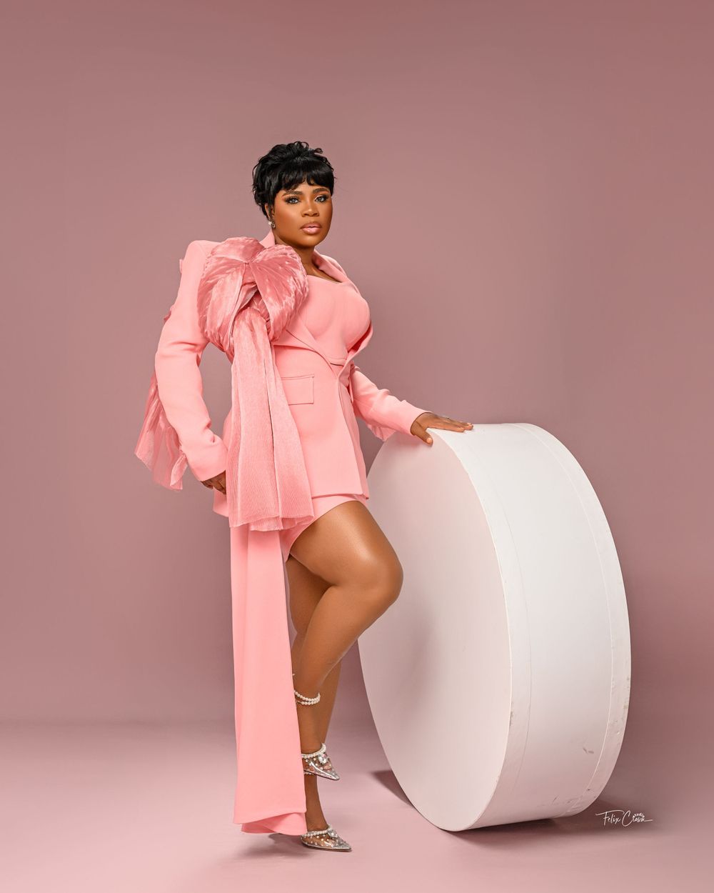 A person in a pink statement dress with dramatic sleeves poses confidently beside a large white cylinder against a mauve backdrop.