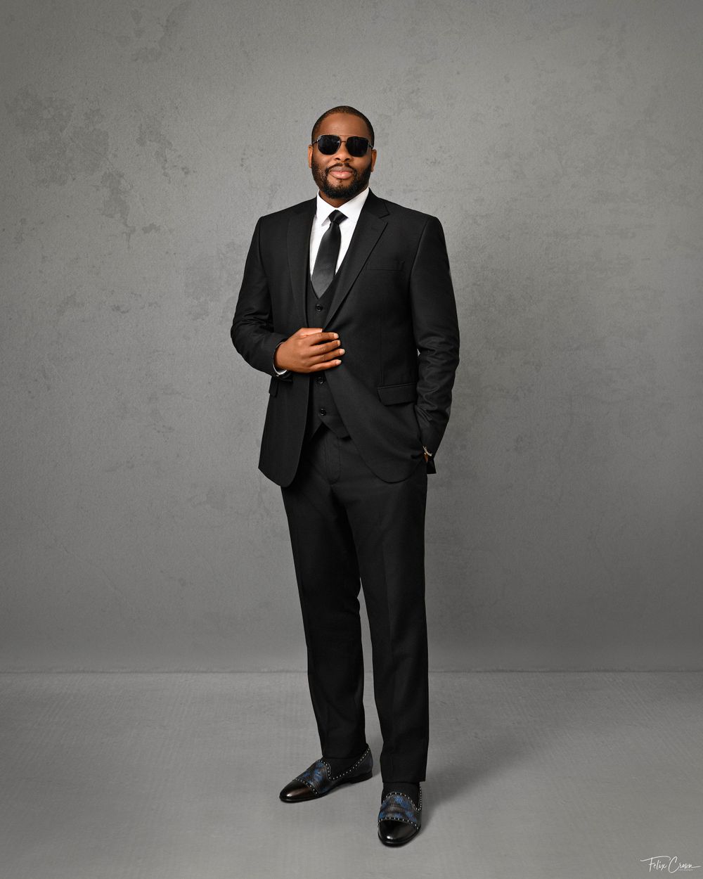 Professional full length portrait of man in black three-piece suit against gray background.