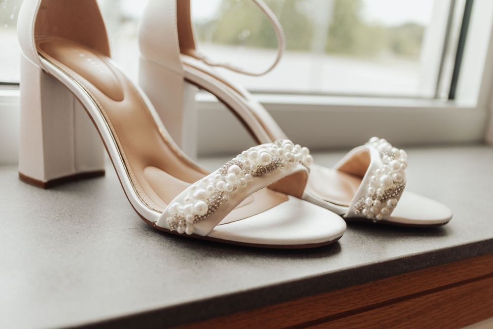 Elegant white wedding heels with beaded straps sitting on a wooden windowsill.