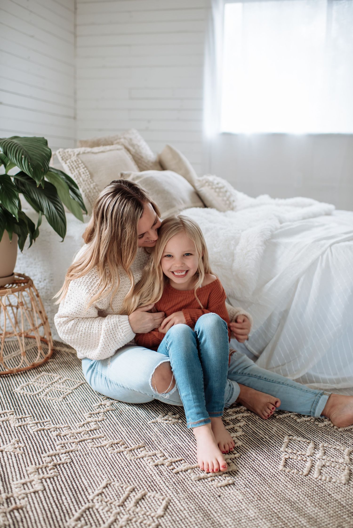 Two people sharing a tender moment while sitting on a patterned rug next to a white bed with houseplants nearby.