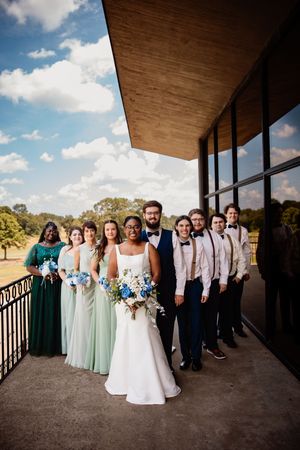Wedding party poses on outdoor terrace with white columns, blue sky and clouds in background. Arkansas Wedding Photographer