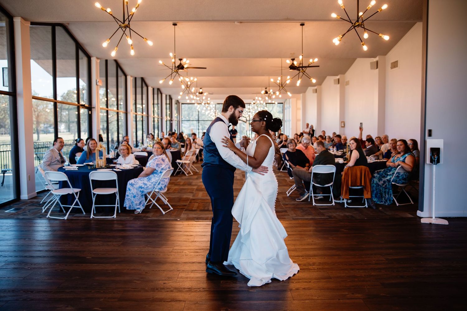 Newlyweds share their first dance on a dark hardwood floor in an elegant venue with floor-to-ceiling windows. Arkansas Wedding Photographer