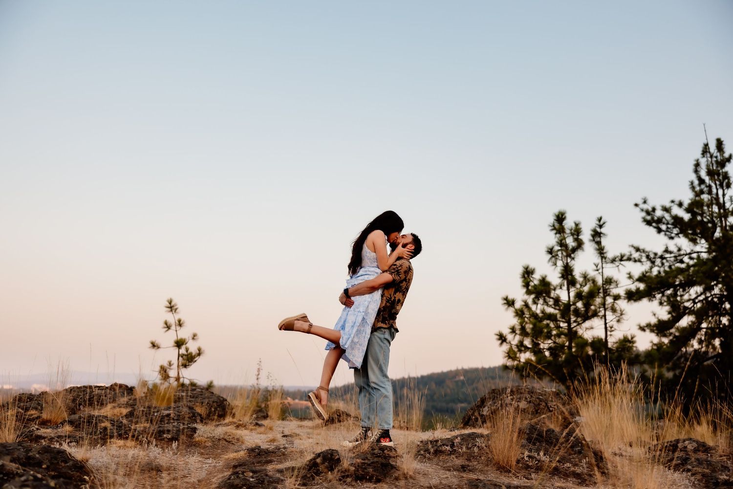 Couple embracing on a scenic hilltop during golden hour with mountains in the background.