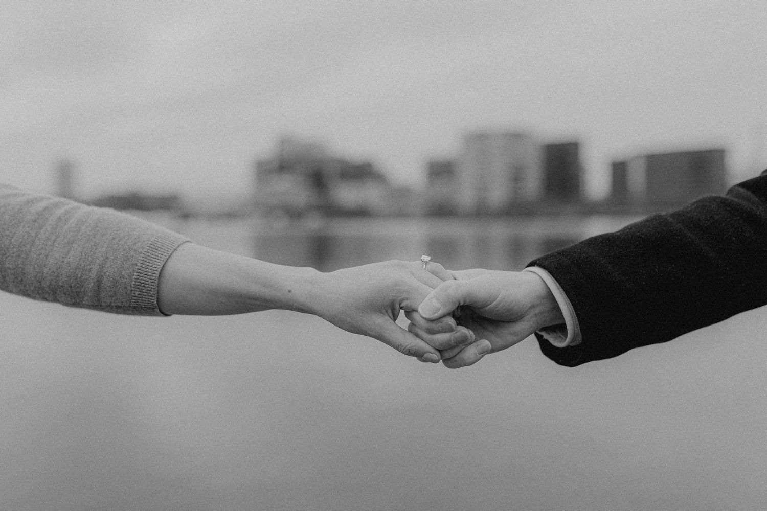 A black and white close-up of two hands reaching to hold each other against a city skyline.