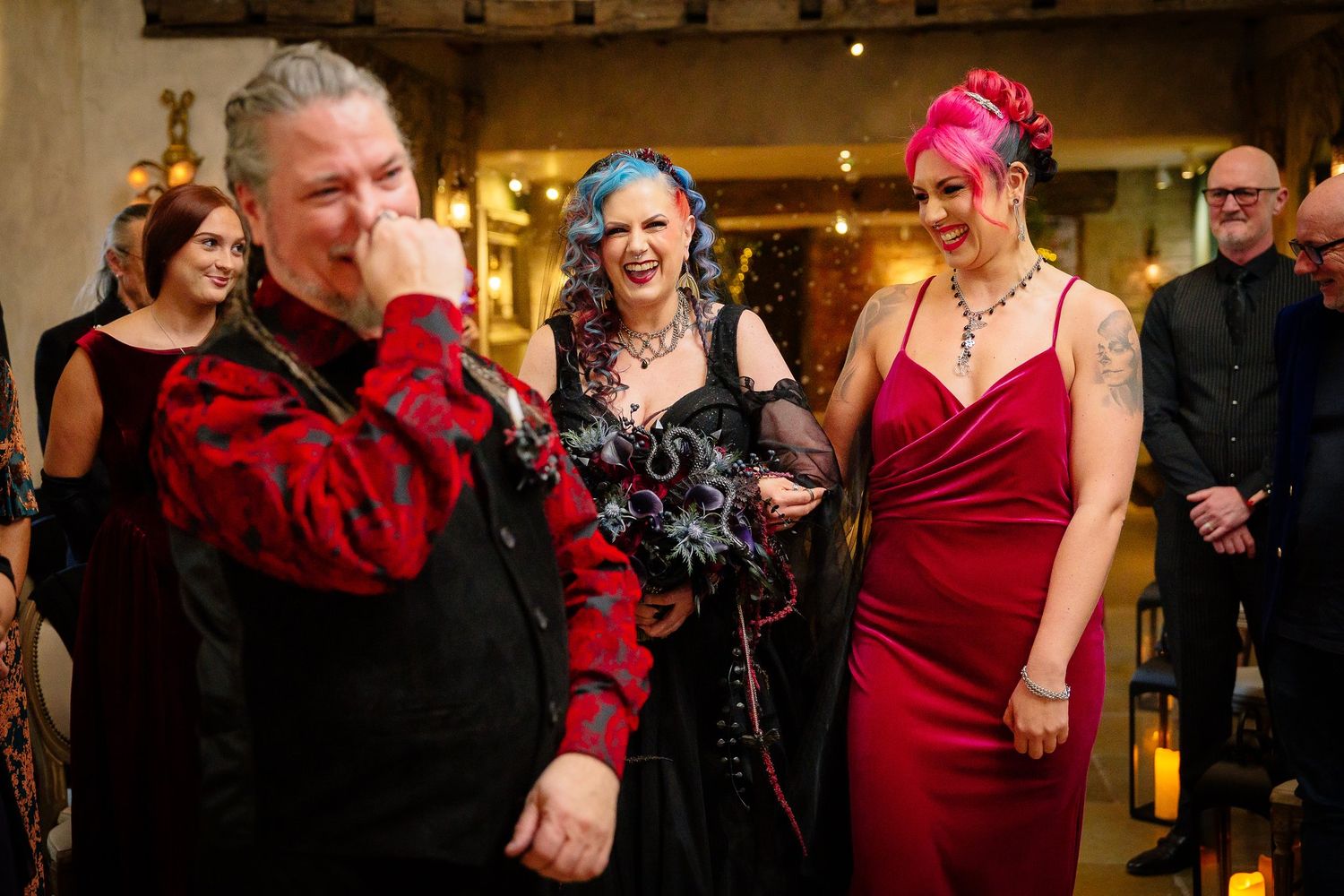 Groom in a red brocade shirt wiping away tears as his gothic bride arrives at their Halloween wedding
