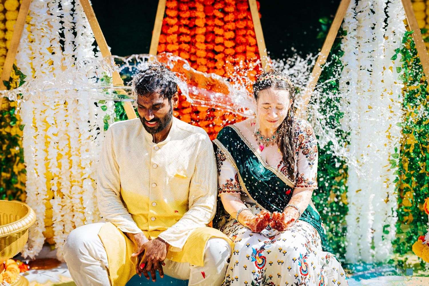 Couple in traditional Indian haldi event attire sit smiling under a floral mandap as water splashes around them during a joyful ceremony.