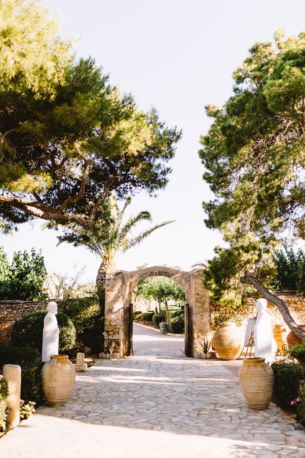 Stone-paved garden pathway leading to a rustic arched gateway, flanked by large terracotta urns, white statues, pine trees and palms.