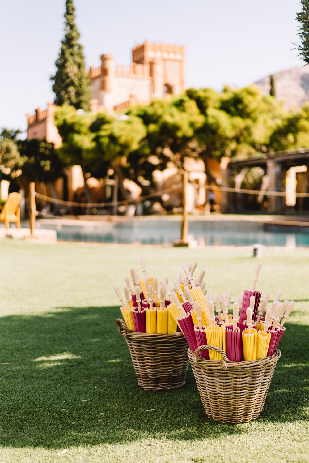 Two wicker baskets filled with pink and yellow paper parasols on a sunny lawn, with a pool and castle-style villa in the background.