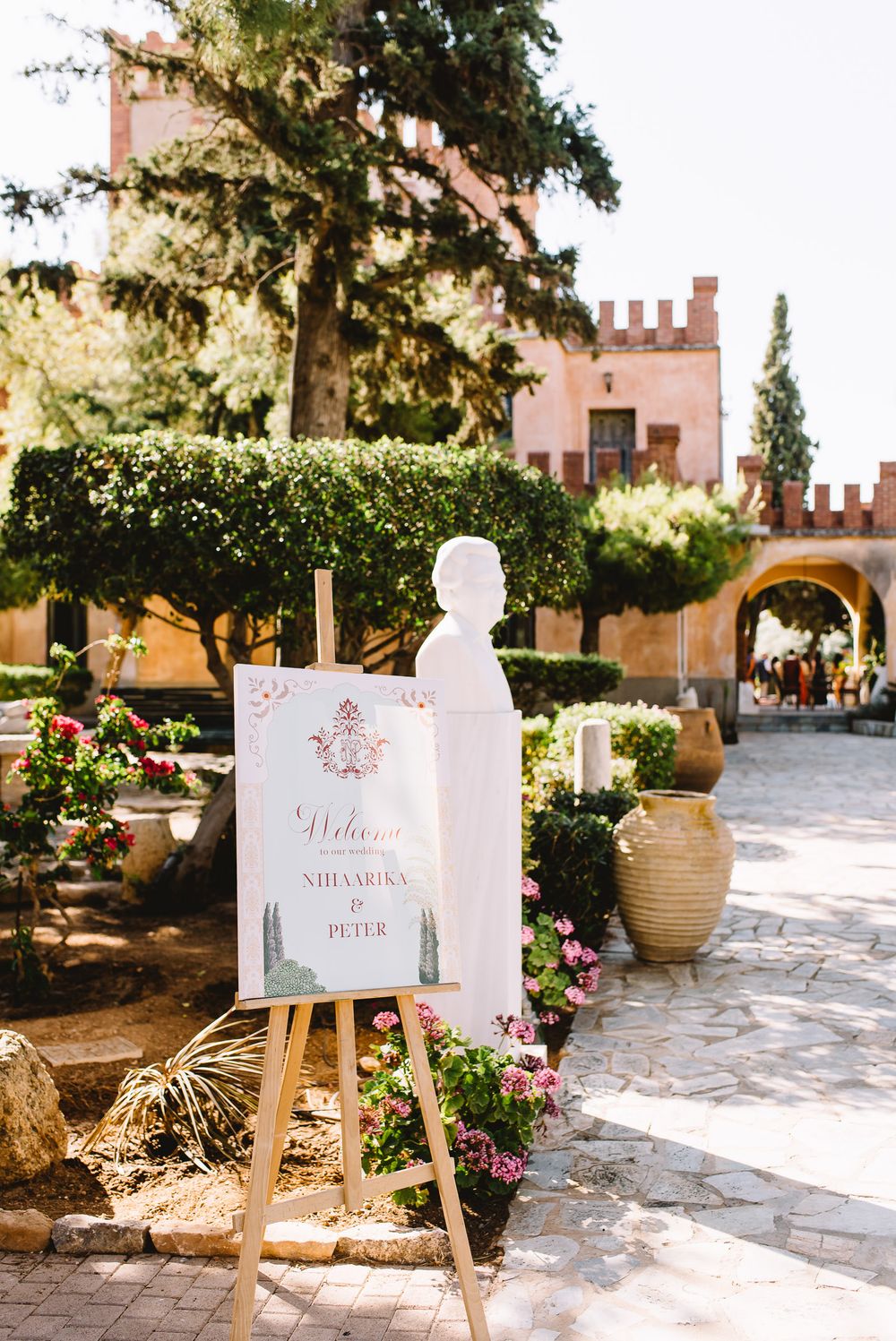 Wedding welcome sign on a wooden easel beside a white bust, pink flowers and a sunlit stone courtyard with a castle-style building.