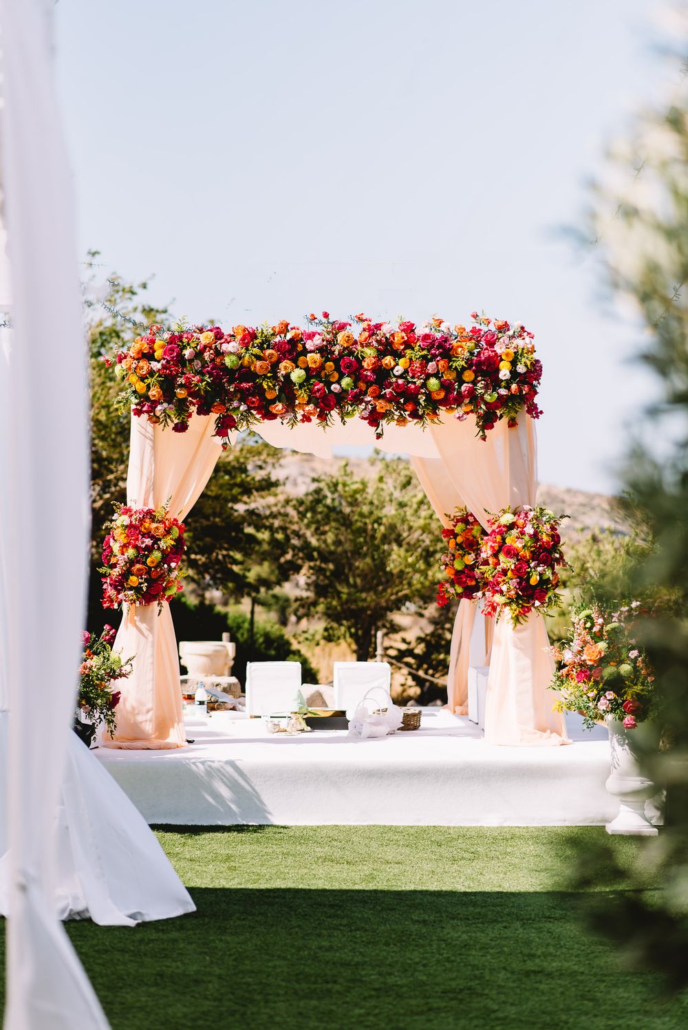 Outdoor wedding mandap with peach draped fabric and a vibrant red, orange and pink floral canopy set on a white platform over green grass.