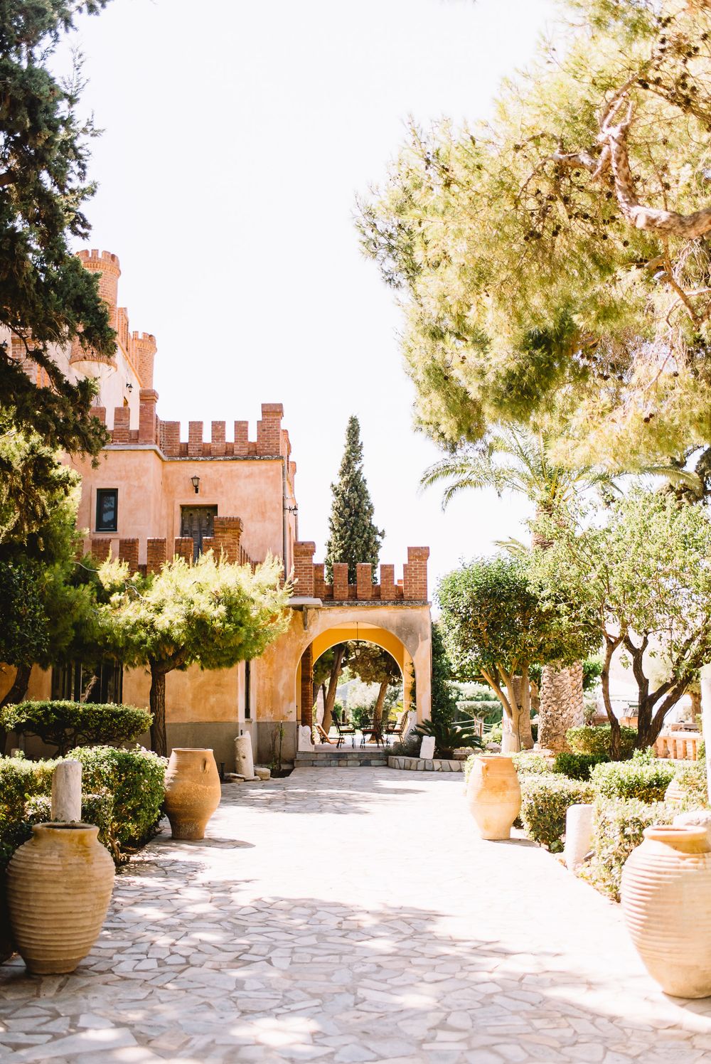 Sunlit stone courtyard with large terracotta urns, olive trees, and a pink crenellated Mediterranean villa with an arched passage.