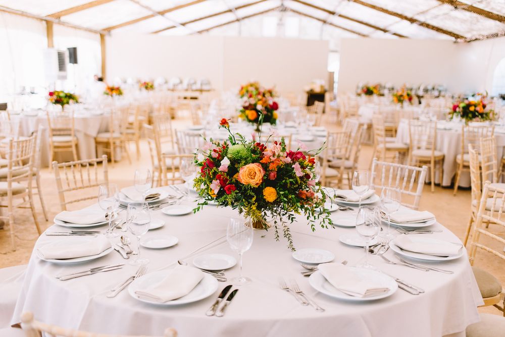Elegant wedding reception table with orange and red floral centerpiece, white linens, and place settings in a bright tented venue.