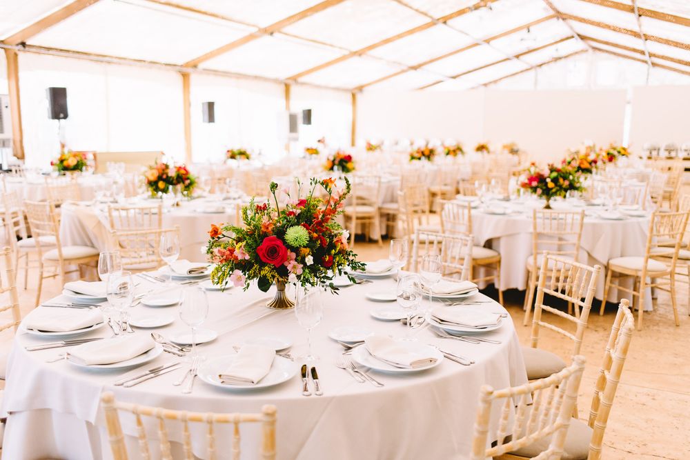 Elegant wedding reception tent with round tables, white linens, place settings, wooden chairs, and red and orange floral centerpieces.