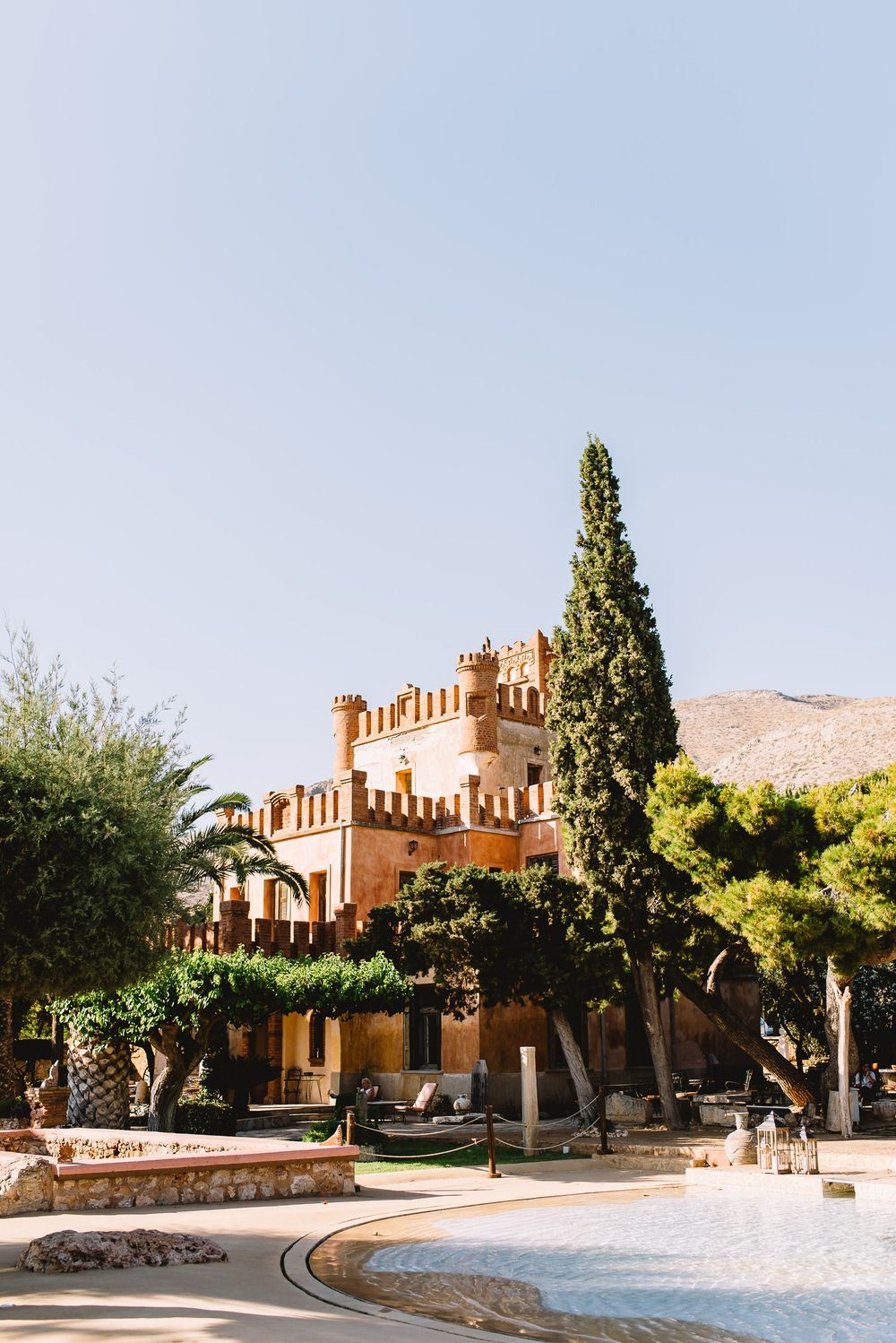 Crenellated terracotta castle with towers, tall cypress and pine trees, and a sunlit pool terrace beneath a clear blue sky and rocky hills.