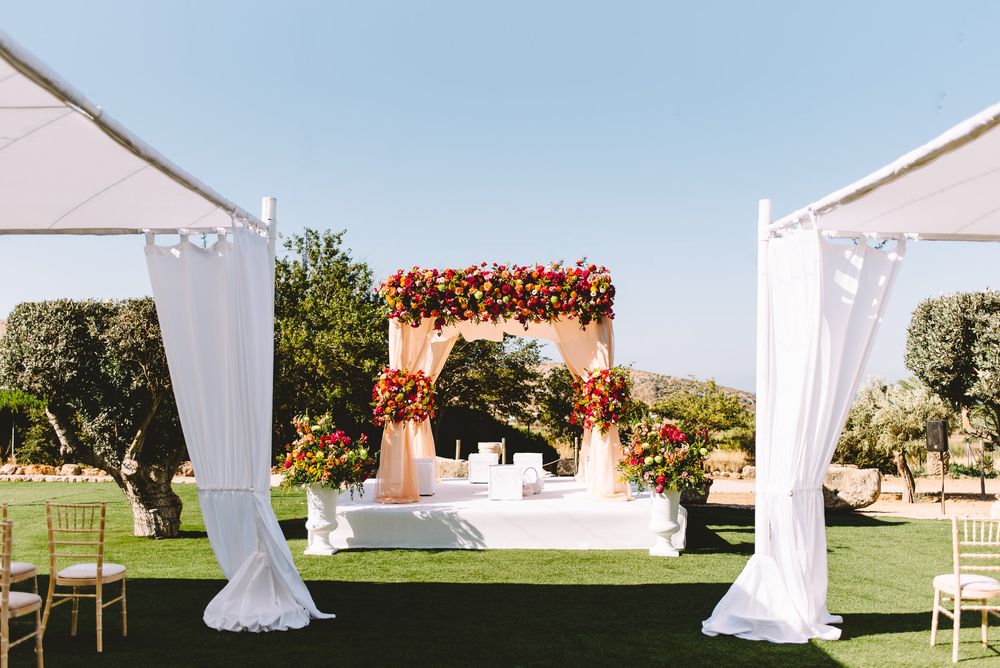 Outdoor wedding ceremony mandap decorated with red and orange flowers on a green lawn, framed by white draped curtains and olive trees.