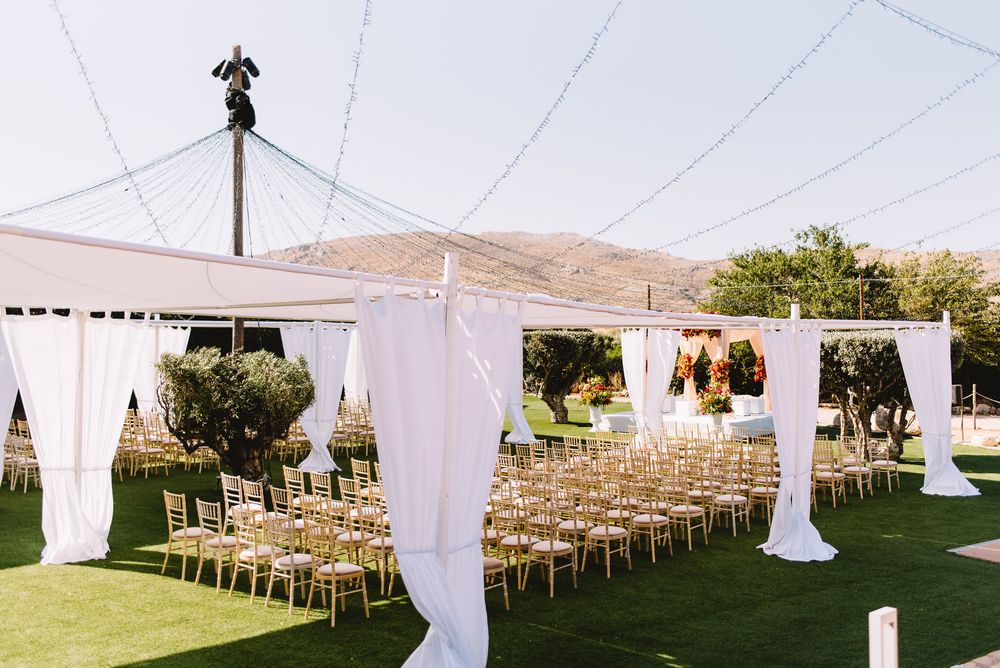 Outdoor wedding ceremony setup with rows of gold chairs under a white draped canopy on a green lawn with hills in the background.
