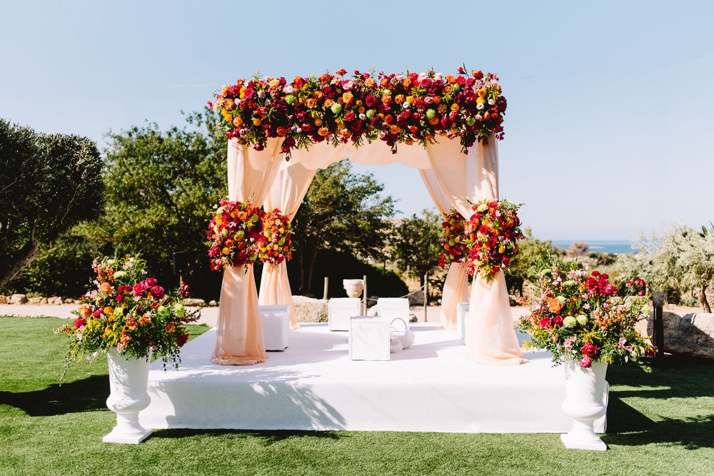 Outdoor wedding ceremony mandap with peach draped fabric and vibrant red, orange and pink floral arrangements on a green lawn near the sea.
