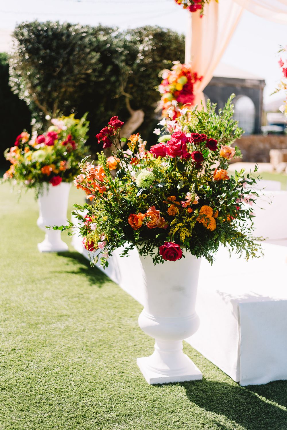 White urns filled with vibrant red, orange and pink flowers and greenery line an outdoor wedding aisle on green grass.