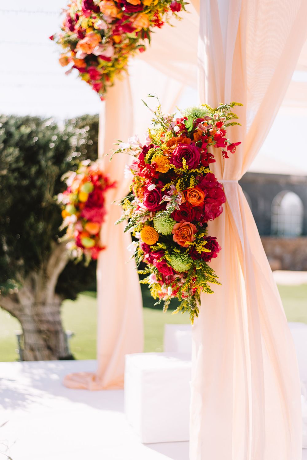 Wedding ceremony canopy with peach draped fabric and vibrant orange, red and green floral arrangements set in an outdoor garden.