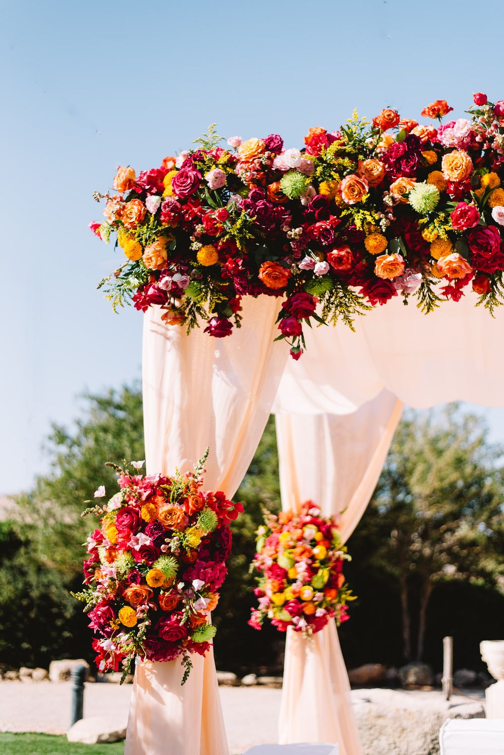 Outdoor wedding arch draped in peach fabric and topped with vibrant red, orange, and pink floral arrangements under a clear blue sky.