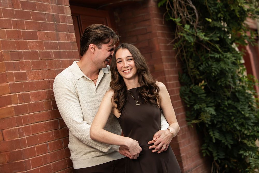A smiling couple embraces against a red brick wall with green ivy, the man whispering close to the woman's ear in a warm, romantic pose.