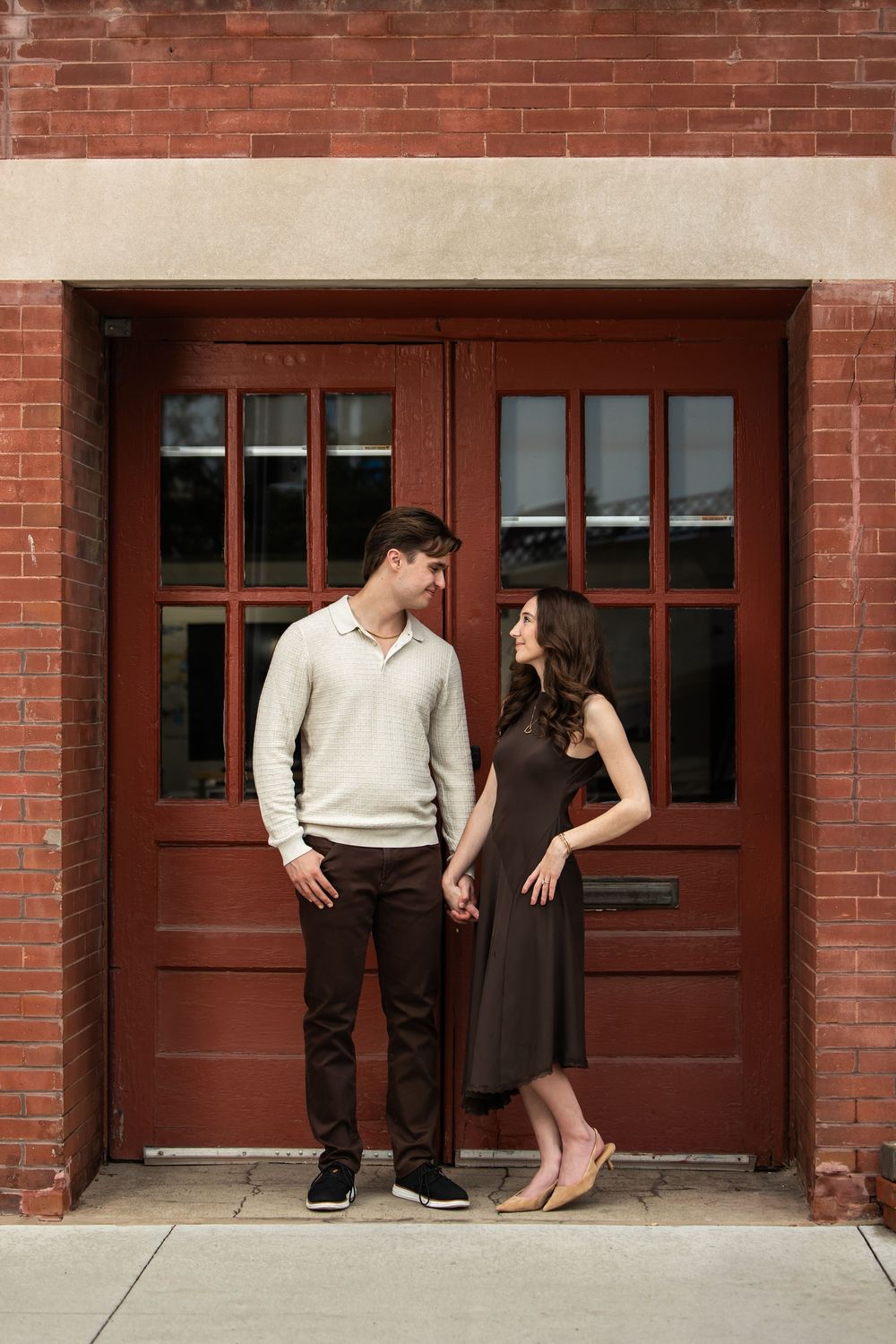 Couple holding hands and smiling at each other in front of a red brick building with rustic double doors during an engagement photo session.