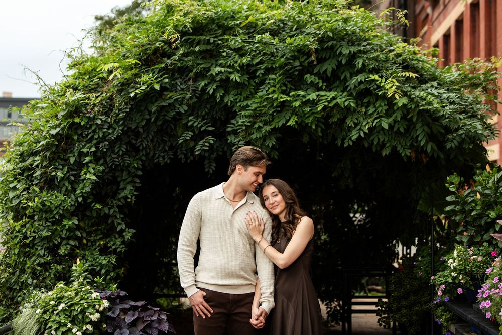 Couple embracing and holding hands in front of a lush green leafy archway during an outdoor engagement photo session.