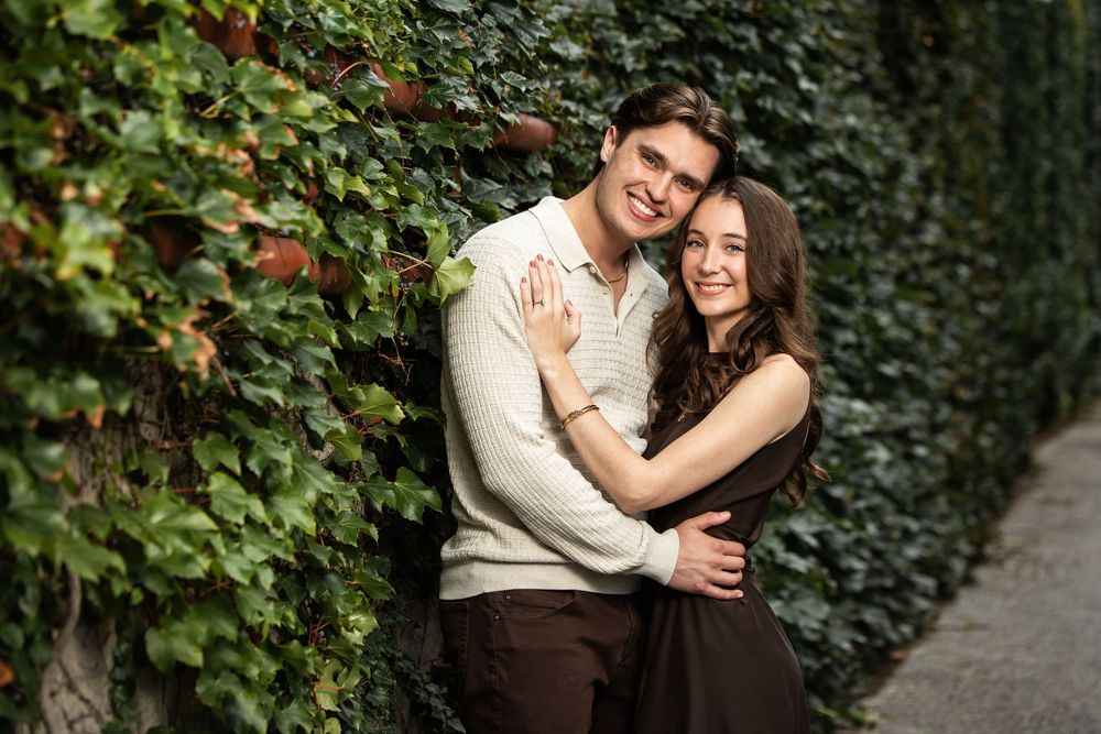 Smiling couple embracing in front of a lush green ivy wall, the woman in a brown dress and the man in a cream sweater.