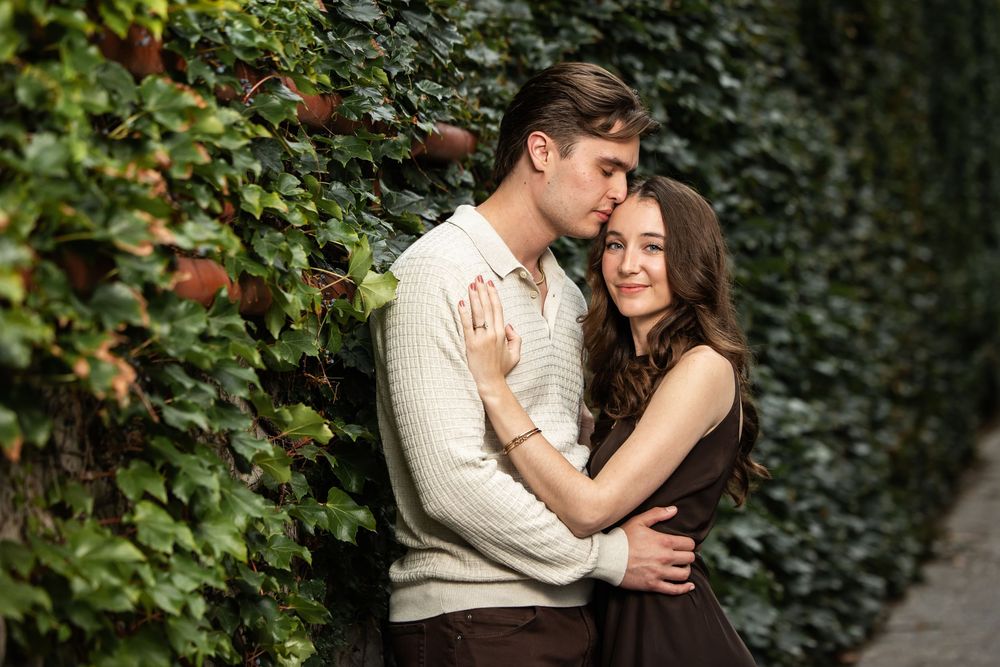 Romantic couple embracing in front of a lush green ivy wall, the man in a cream sweater kissing the smiling woman's forehead.