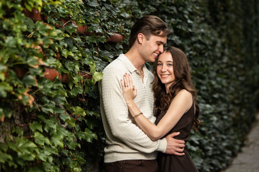 Smiling couple embracing in a romantic engagement-style pose against a lush green ivy wall, the man kissing her forehead.