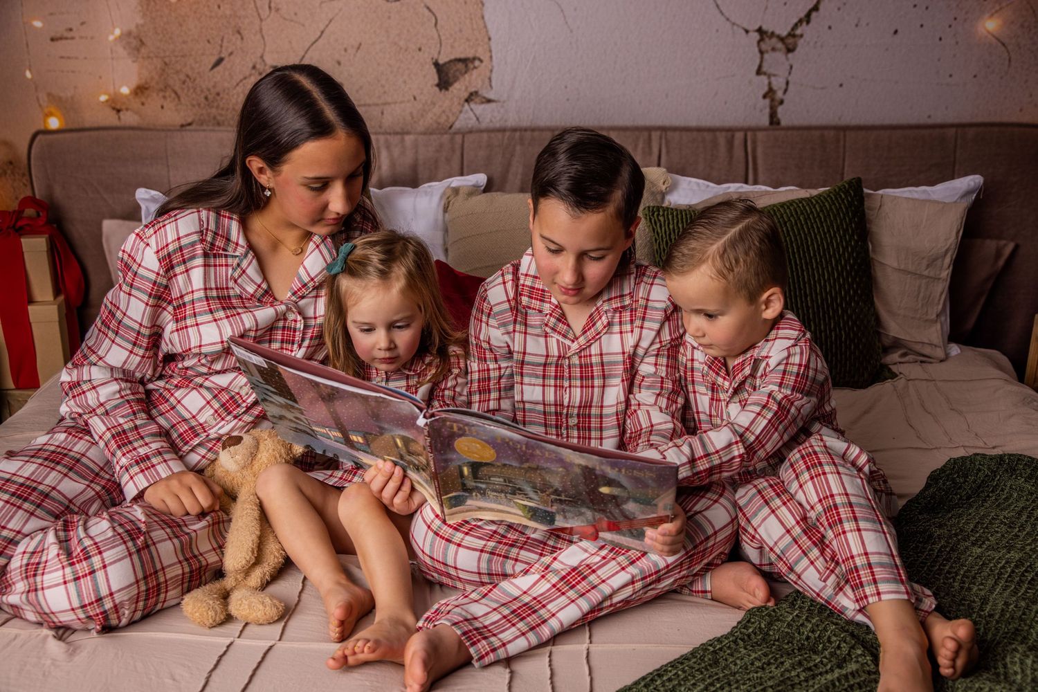 Mother and three children in matching red plaid pajamas sit on a bed reading a picture book together beside a teddy bear.