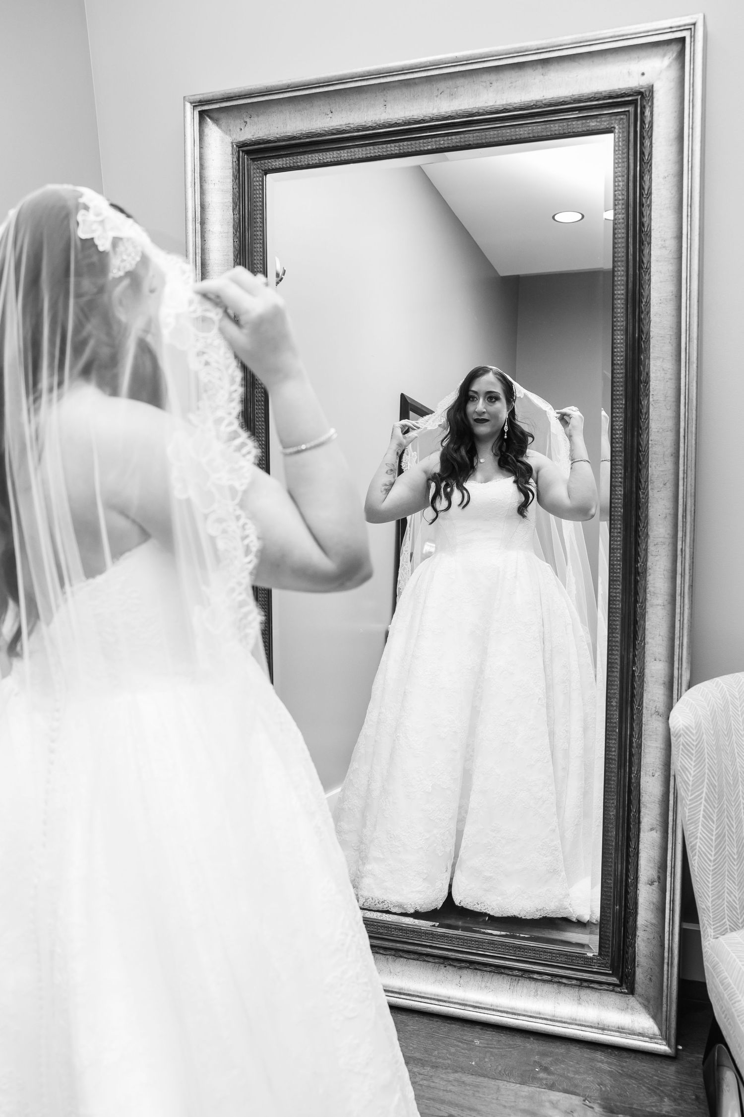 Black and white photo of a bride in a lace wedding gown adjusting her veil while admiring her reflection in a large framed mirror.