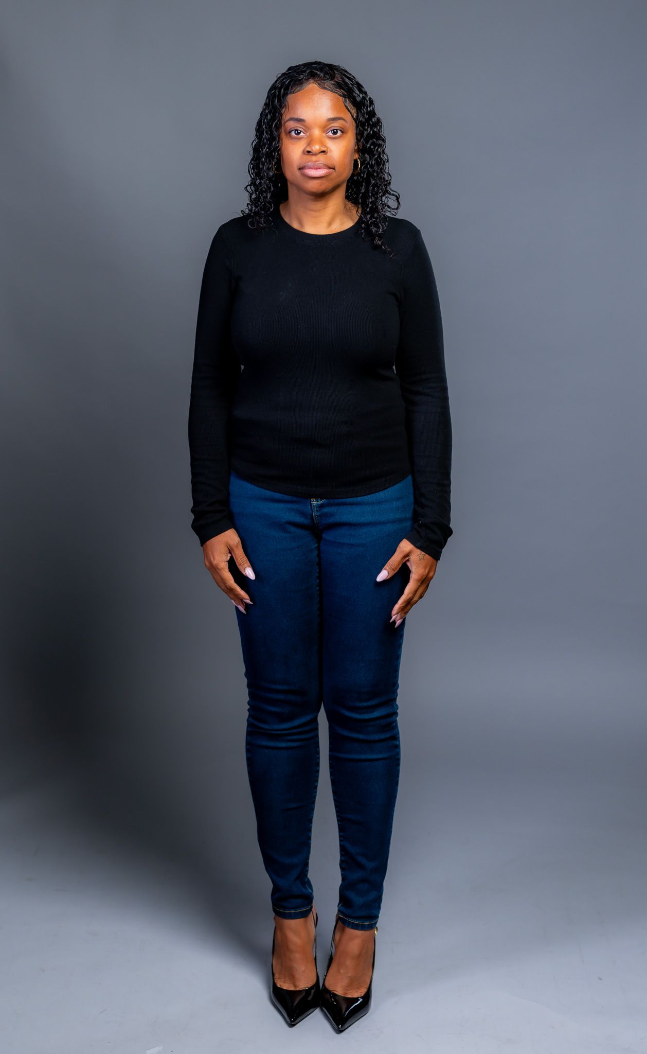 Full-length model portrait in a black sweater, blue jeans and black heels against a gray studio background.