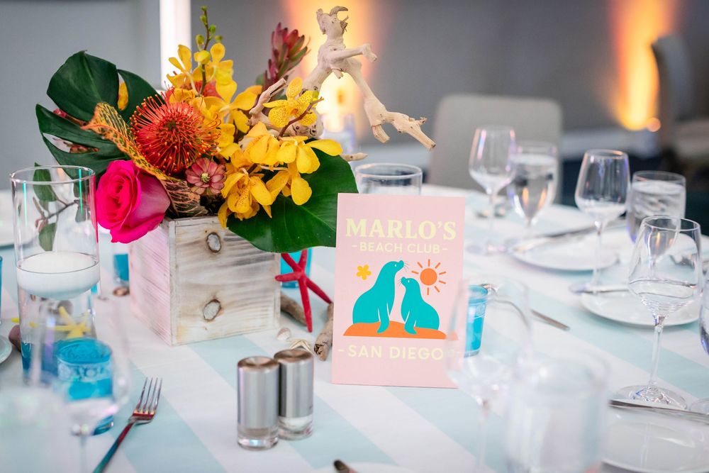 Tropical flower centerpiece in a wooden box beside a Marlo's birthday sign with teal dolphins on a striped table set with glassware.