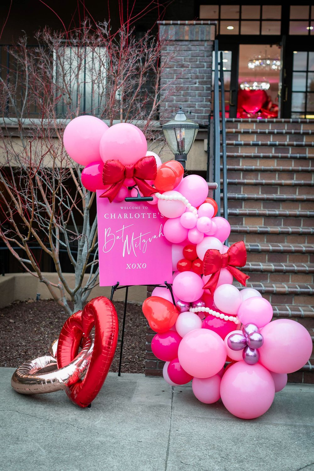 Pink, red and white balloon garland with bows and heart foil balloons beside a Charlotte welcome sign at a stairway entrance.