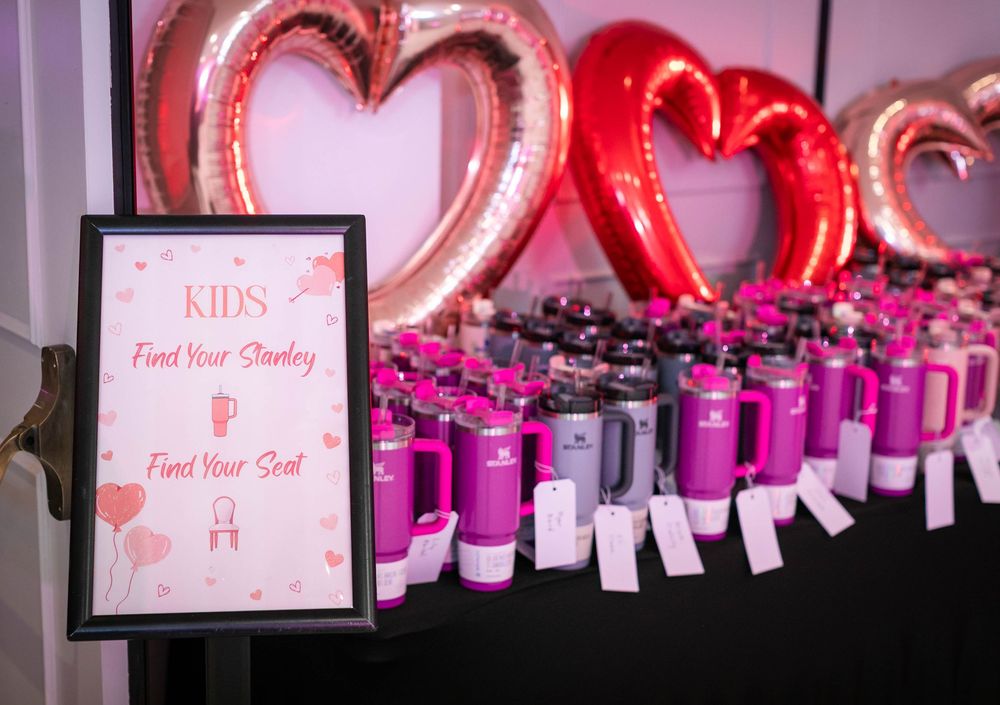 Pink and gray tumblers with name tags on a black table beneath heart-shaped balloons, with a sign reading Find Your Stanley, Find Your Seat.