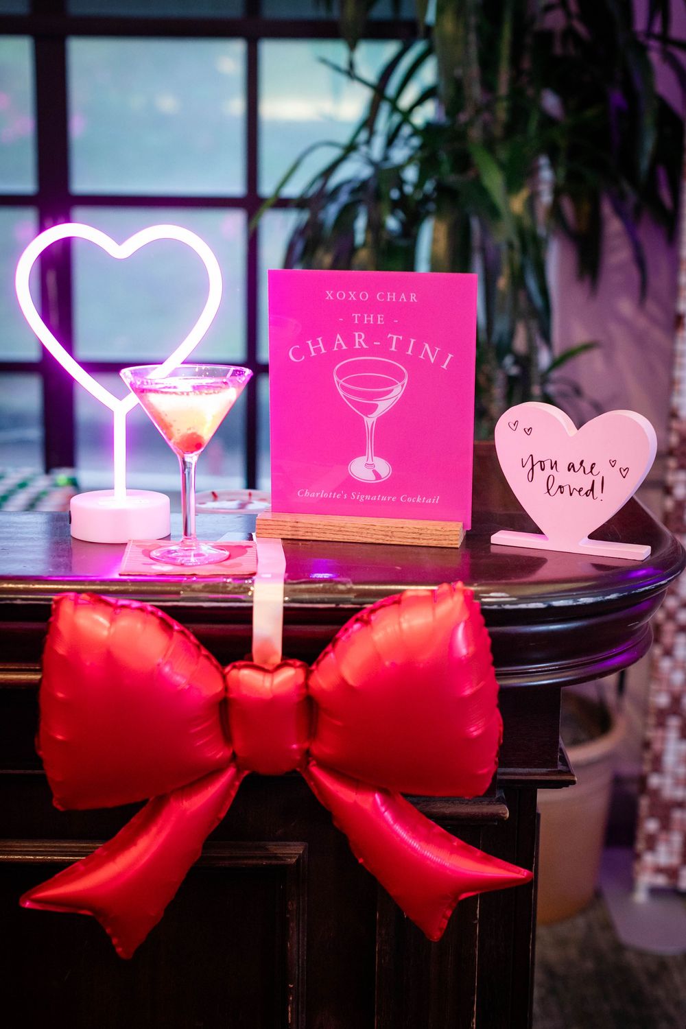 Pink cocktail in a martini glass beside a glowing neon heart, a Char-Tini menu card, a You are loved sign, and a red bow on a bar.