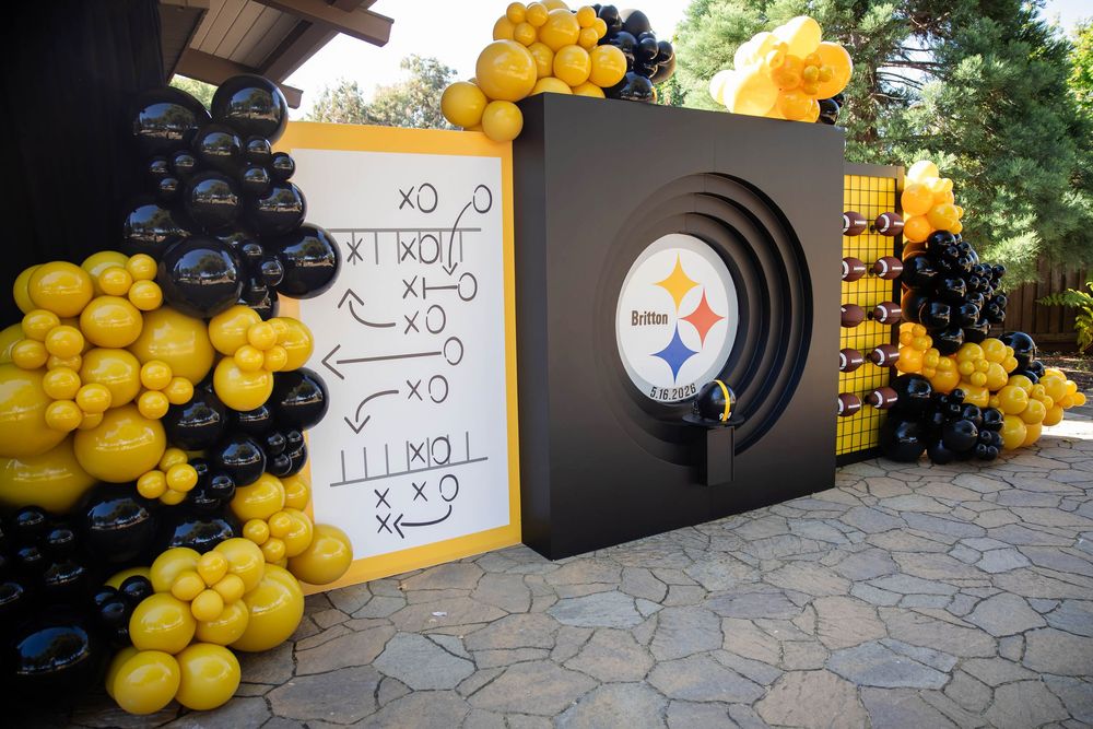 Black and gold balloon garland with a Pittsburgh Steelers logo display and football play diagram board at a themed outdoor party.