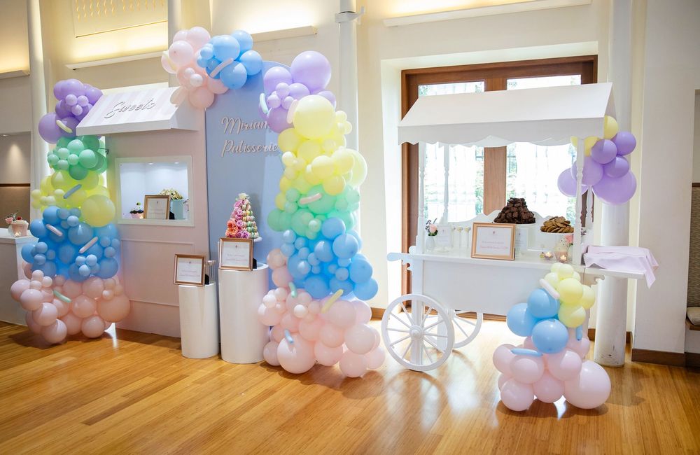 Pastel balloon garlands in blue, pink, yellow and lavender frame a white dessert cart and candy stand at a party venue.