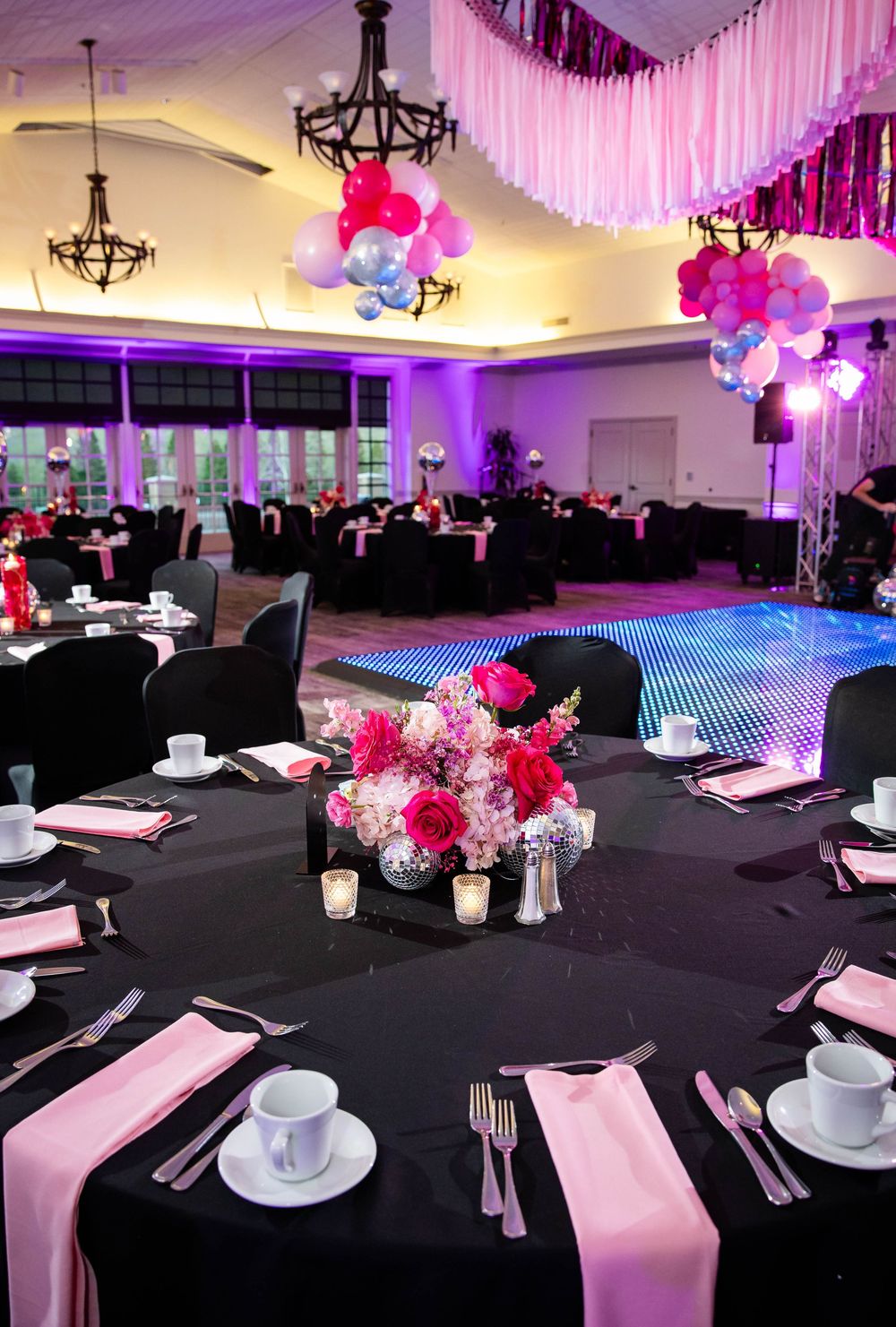 Elegant banquet hall with black tablecloths, pink napkins, floral centerpieces, balloon clusters, and a lit LED dance floor.