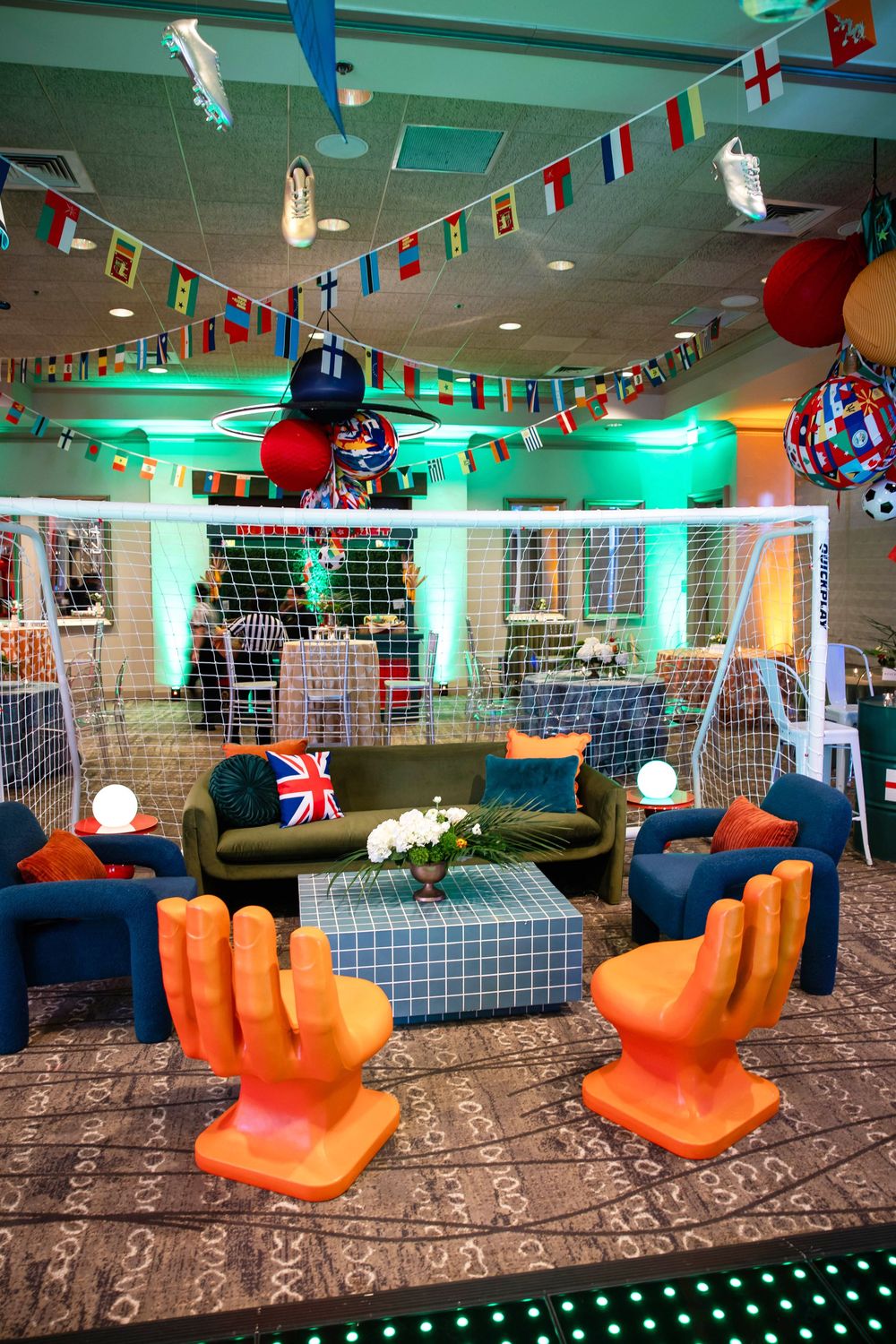 Soccer-themed event lounge with orange hand-shaped chairs, a Union Jack pillow, international flag bunting and a goal net backdrop.