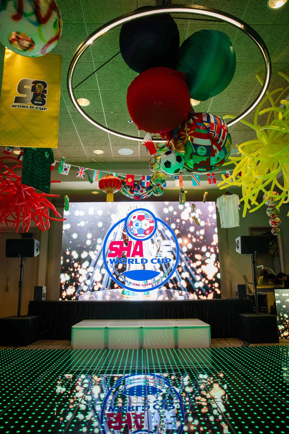 World Cup themed event stage with LED screen, soccer ball balloons, international flags, and a lit dance floor under a ring chandelier.