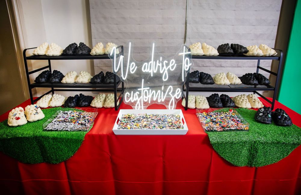 Custom sneaker bar display with a neon sign reading "We advise to customize," shoe racks, and a red table with turf-covered side tables.