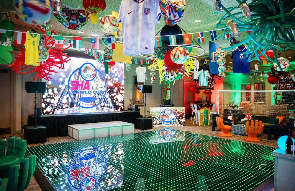 Festive event space with a glowing LED dance floor, stage with video screen, and sports jerseys and flags hanging from the ceiling.