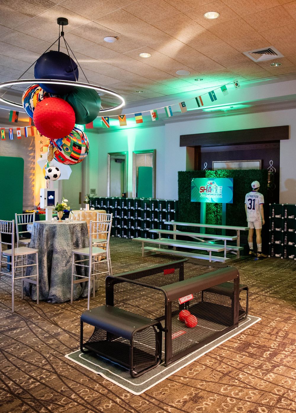 Sports-themed event space with a sports-ball chandelier, flag bunting, a green hedge wall, benches, and a draped table with chairs.