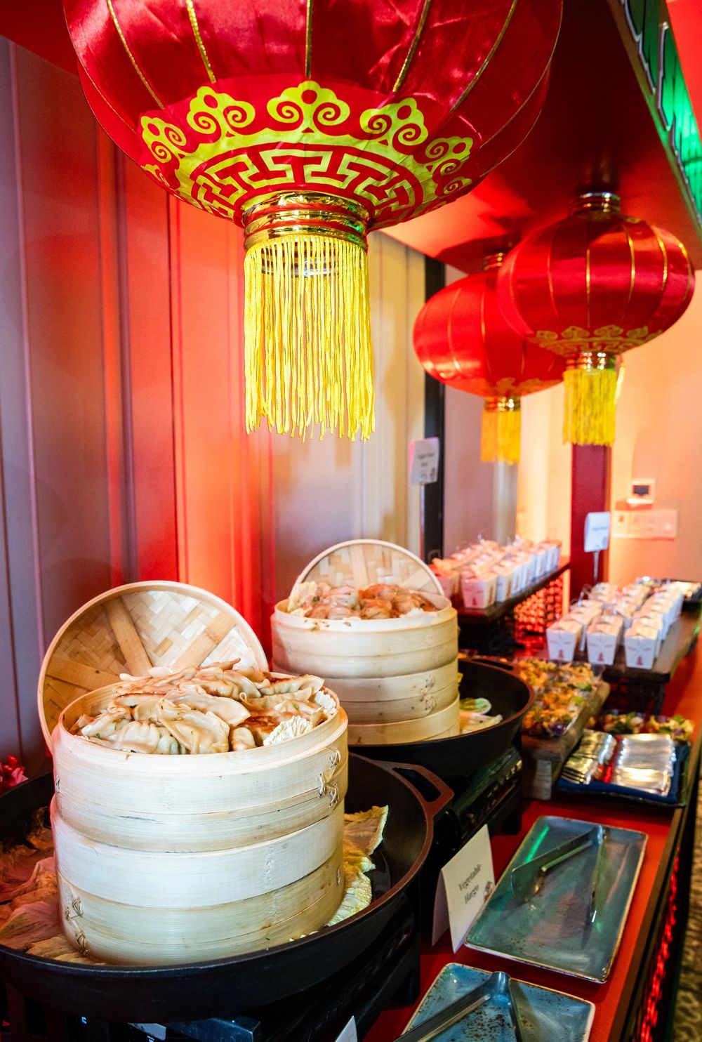 Bamboo steamers filled with dumplings on a buffet counter beneath red Chinese lanterns with gold tassels, with serving trays nearby.