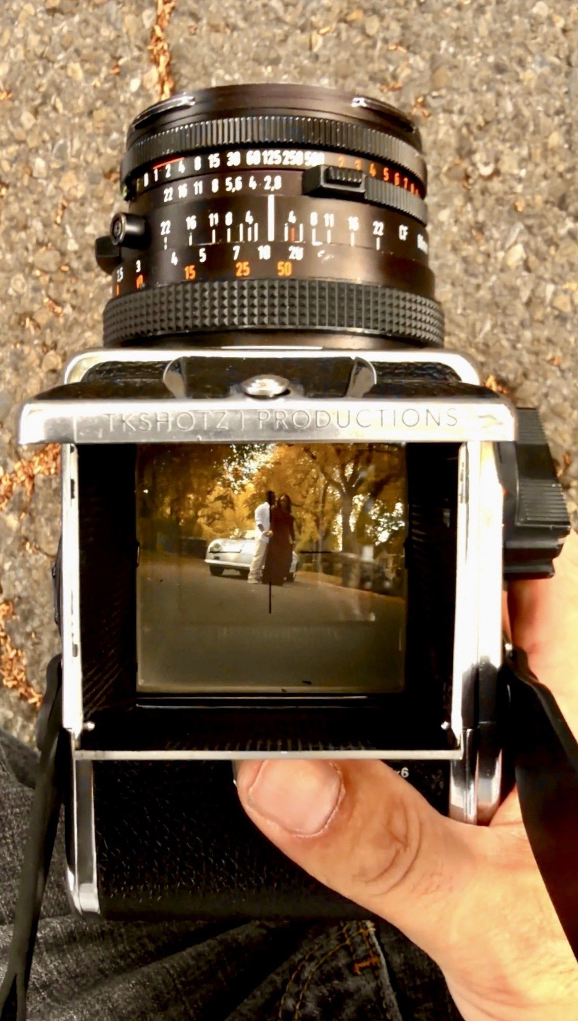 Hand holding a vintage medium format film camera with open waist-level viewfinder showing a reflected autumn street scene.