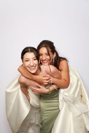 Two women in formal dresses embrace and laugh together against a white background.