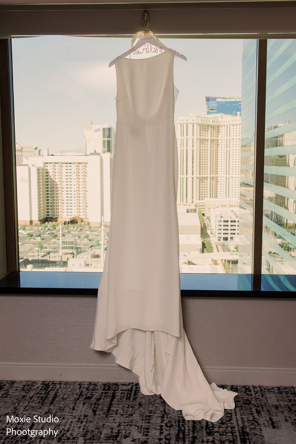 Elegant white sleeveless wedding dress with a flowing train hangs in a hotel window overlooking a sunny city skyline.